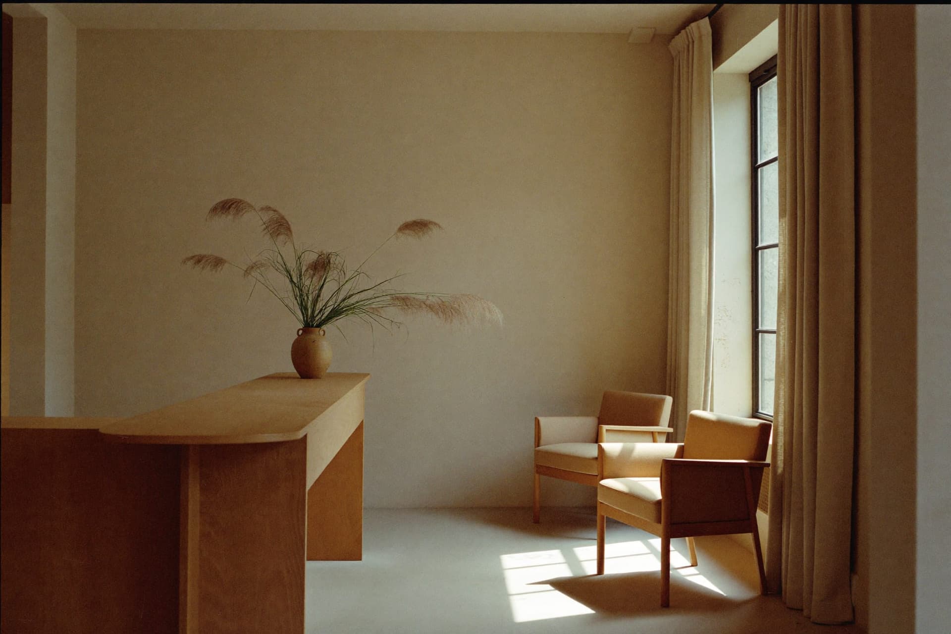 A quiet consultation room with a light oak desk, two linen chairs and a sunlit window