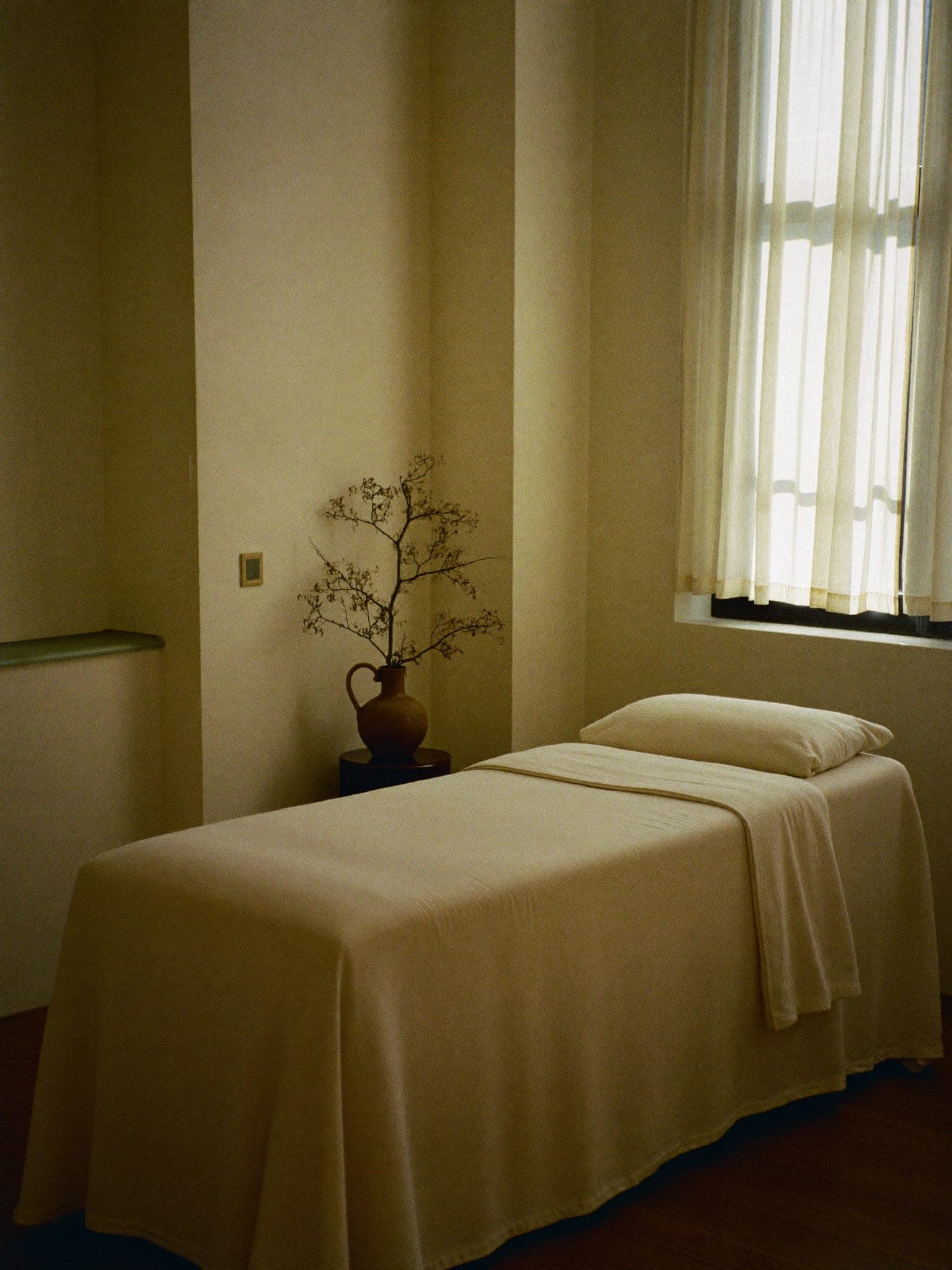 A calm, empty treatment room with a linen-draped bed beside a curtained window in afternoon light